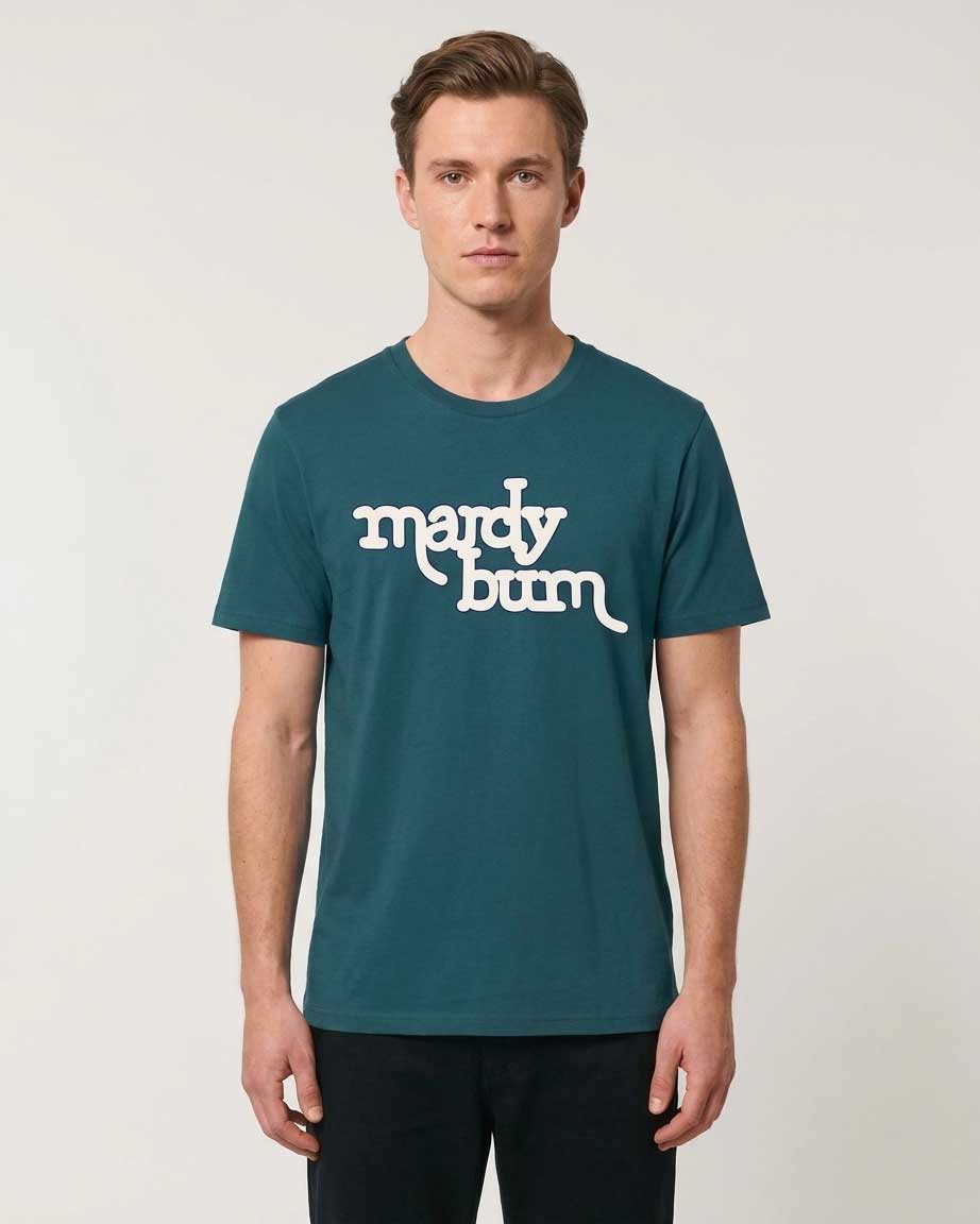 A man wearing a dark teal t-shirt with a white graphic text print that reads 'mardy bum' on the front, paired with black pants, standing against a light gray studio background.